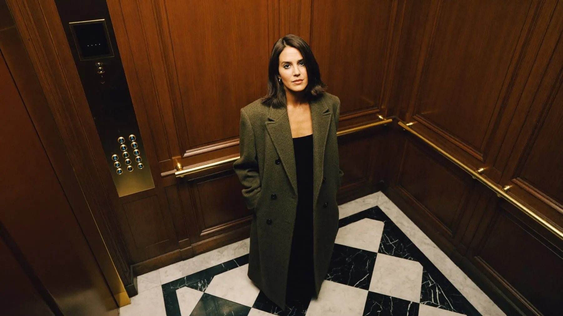 A woman in an olive coat standing in a walnut-and-brass elevator, looking up